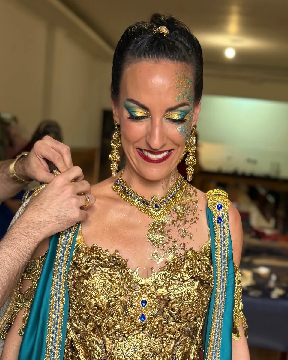 Una festera sonríe con los ojos maquillados en turquesa y oro, labios granates y pan de oro en el cuello mientras le abrochan el traje