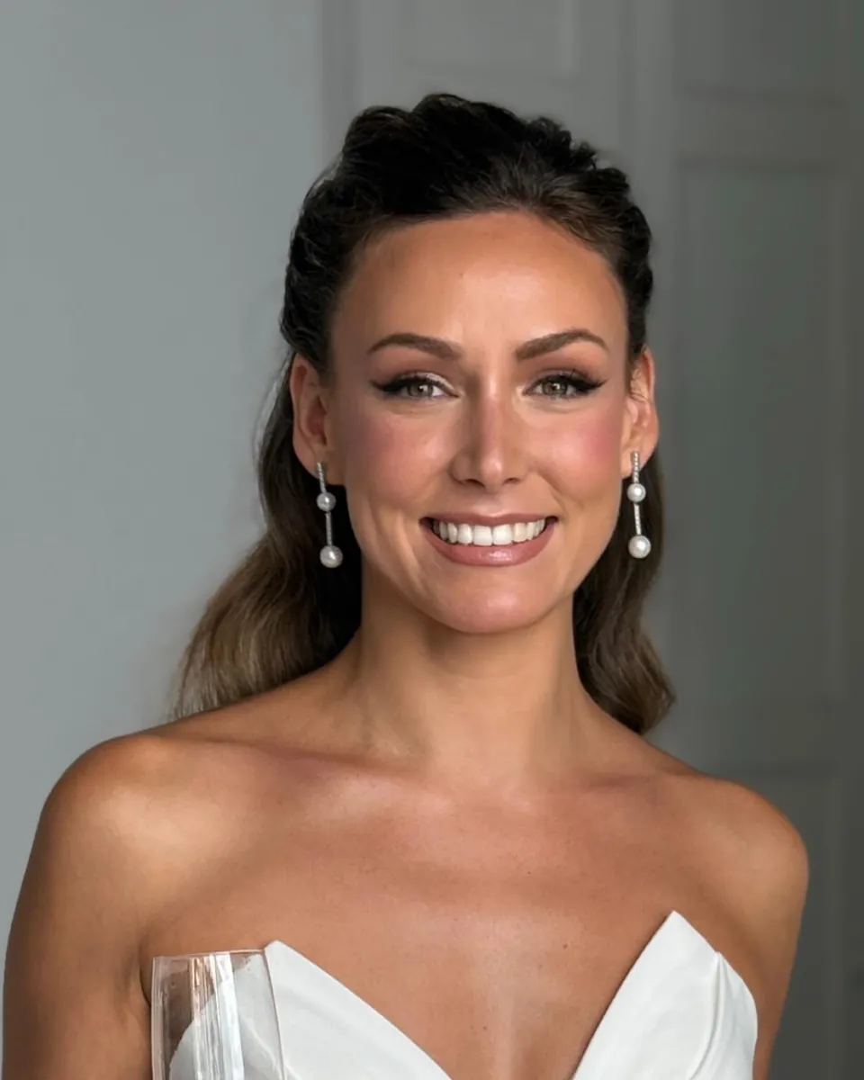 Novia sonriendo con una copa en la mano, pendientes de perlas y el pelo recogido a medias