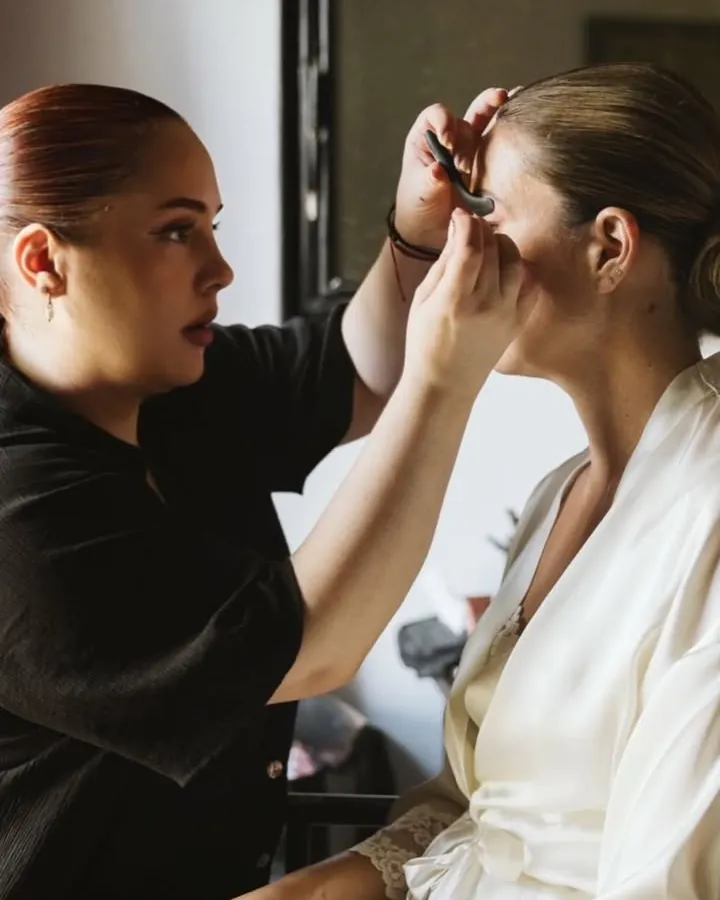 La maquilladora perfilando los ojos a una novia en bata de satén blanca