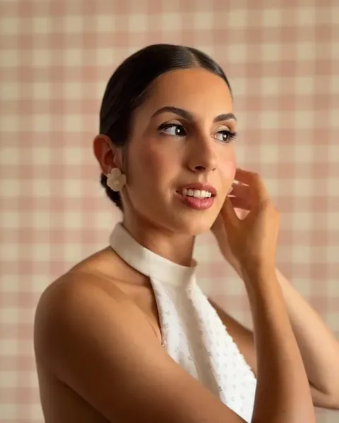 Mujer de perfil con vestido blanco de cuello halter, tocándose el pendiente ante un fondo de cuadros rosas