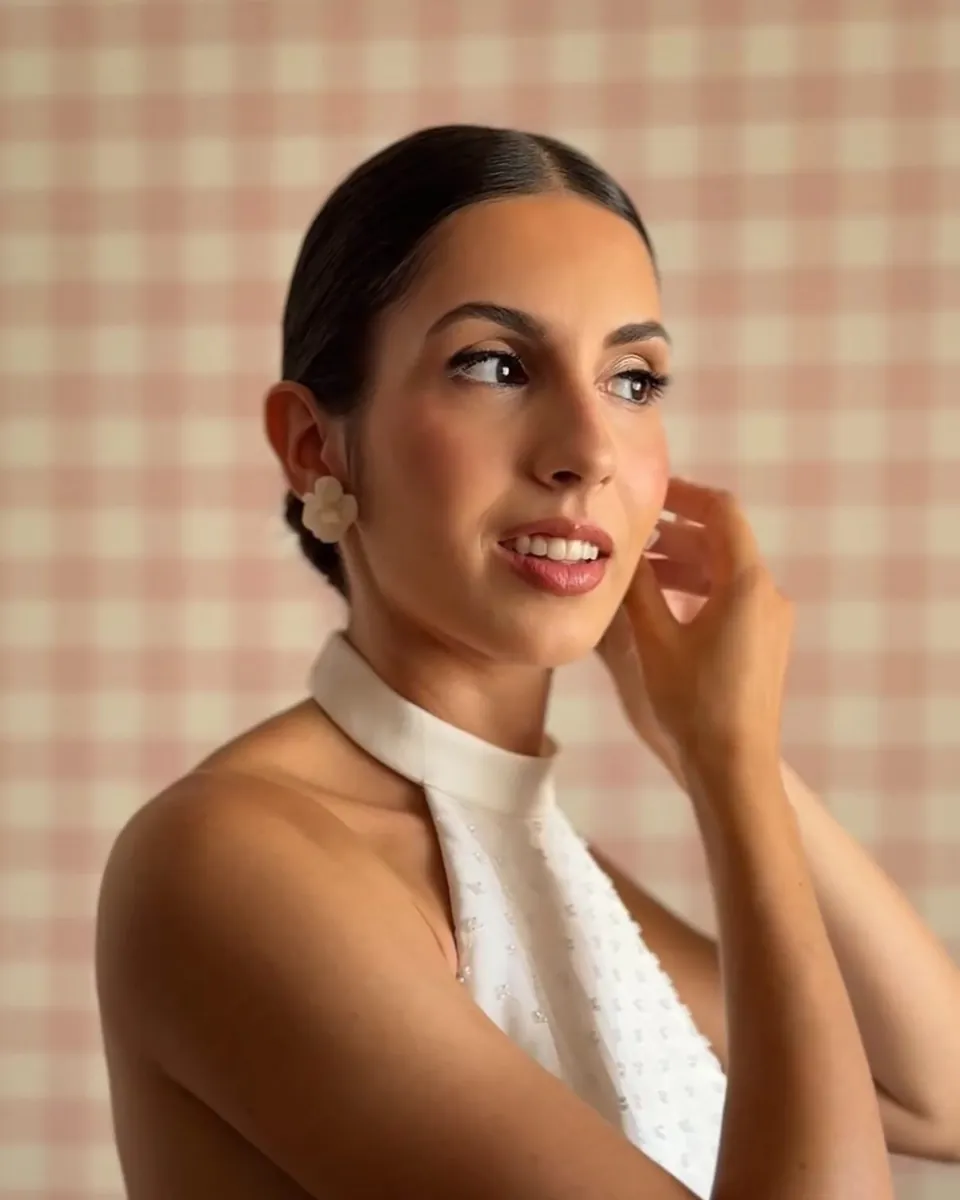 Mujer de perfil con vestido blanco de cuello halter, tocándose el pendiente ante un fondo de cuadros rosas