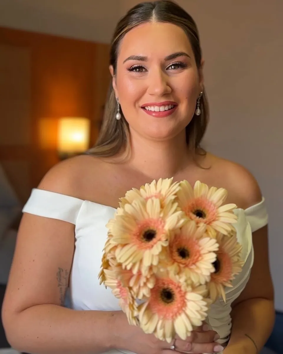 Novia sonriendo con un ramo de gerberas color melocotón y una banda en el vestido