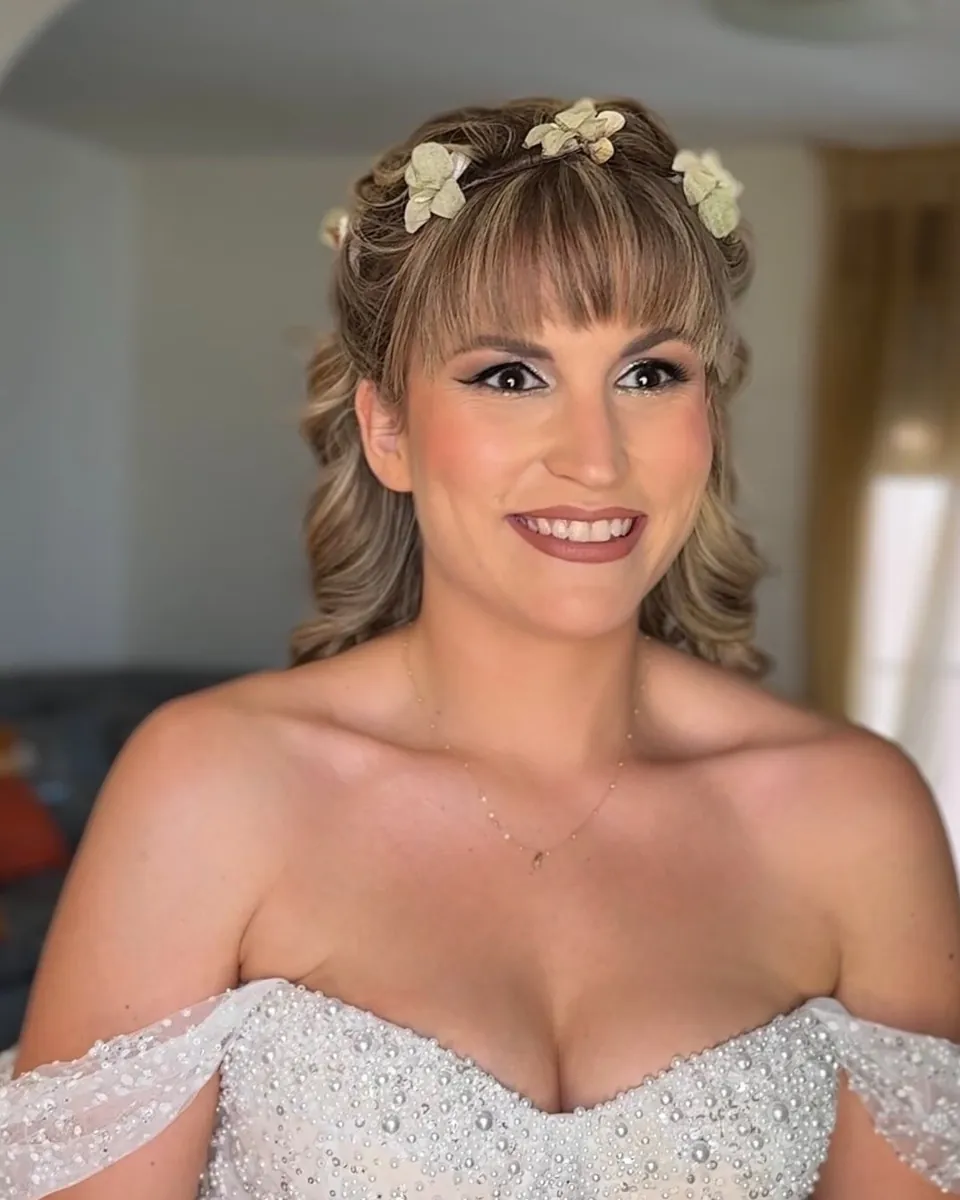 Novia con flequillo, flores en el pelo y vestido de hombros caídos con brillo