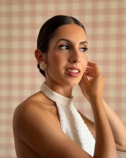 Novia de perfil con vestido de cuello halter, tocándose el pendiente ante un fondo de cuadros rosas
