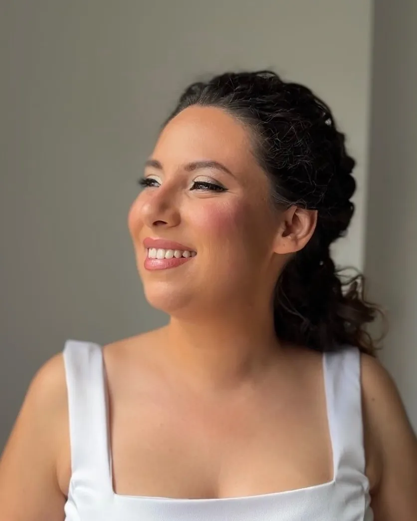 Novia con el pelo recogido en trenzas y vestido de tirantes, sonriendo de perfil junto a una ventana